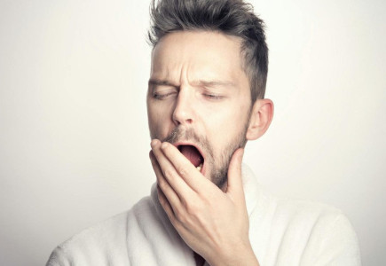 Close-up of a bearded man yawning with hand on mouth against a white background.