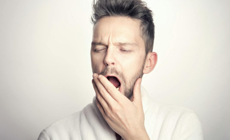 Close-up of a bearded man yawning with hand on mouth against a white background.