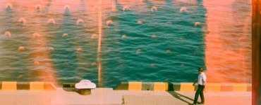 Man walks along a sunny waterfront with teal water.