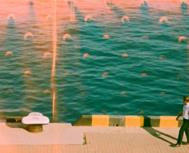 Man walks along a sunny waterfront with teal water.