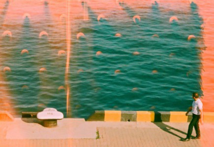 Man walks along a sunny waterfront with teal water.