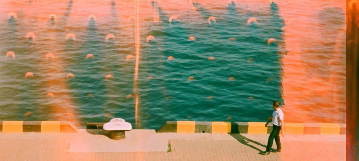 Man walks along a sunny waterfront with teal water.