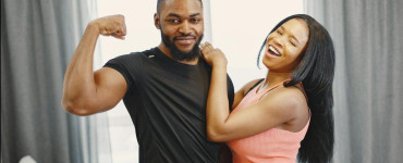 Joyful couple indoors with man flexing bicep and woman laughing, conveying happiness and strength.