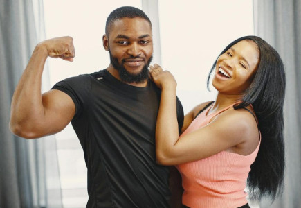Joyful couple indoors with man flexing bicep and woman laughing, conveying happiness and strength.