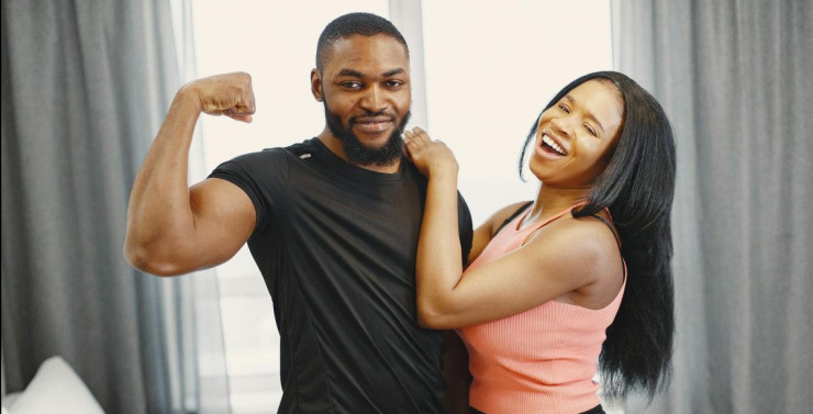 Joyful couple indoors with man flexing bicep and woman laughing, conveying happiness and strength.