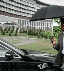 African American man in a suit walking by a car under an umbrella on a rainy day.