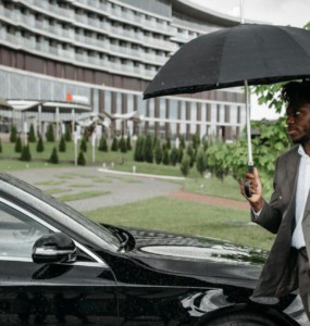 African American man in a suit walking by a car under an umbrella on a rainy day.