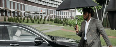 African American man in a suit walking by a car under an umbrella on a rainy day.