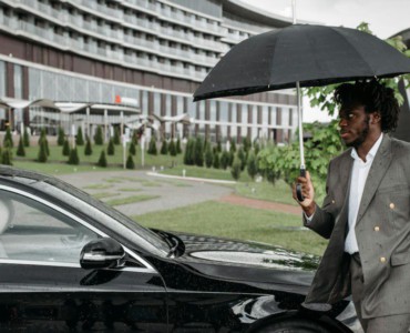 African American man in a suit walking by a car under an umbrella on a rainy day.