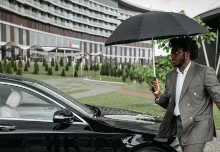 African American man in a suit walking by a car under an umbrella on a rainy day.