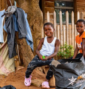 Two smiling children in front of a wooden house.