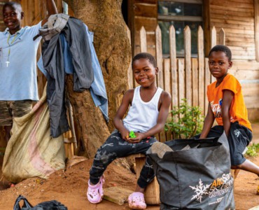 Two smiling children in front of a wooden house.